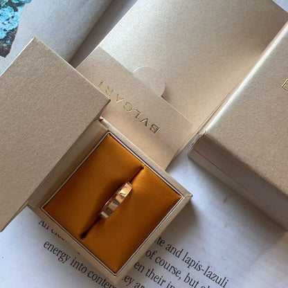 A gold ring presented in an open beige Bulgari jewelry box with a gold interior, photographed on top of printed pages.