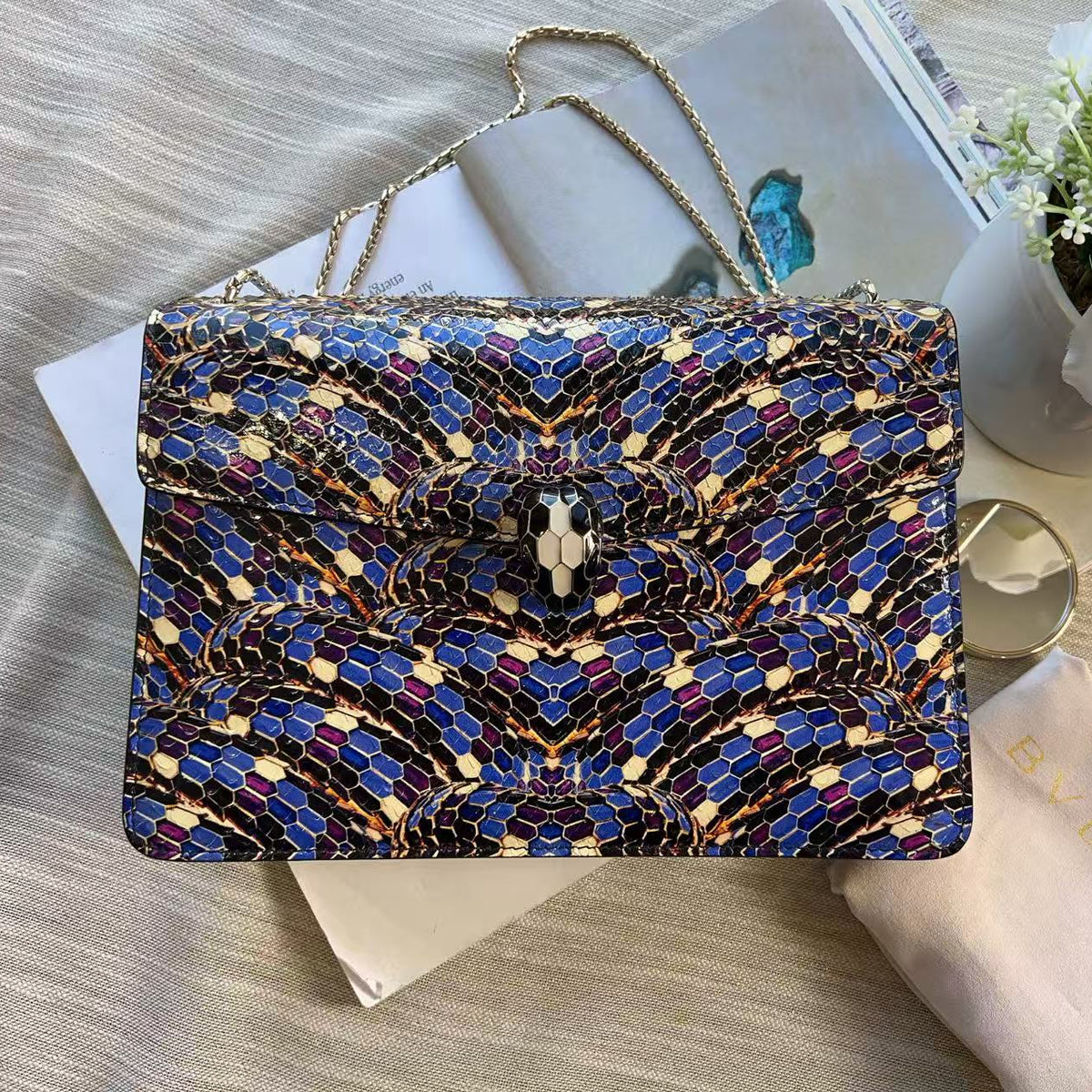 A rectangular clutch bag with a blue, gold, and black snakeskin pattern, styled on top of a white book with a gold chain strap, next to a white Bvlgari dust bag and white flowers.