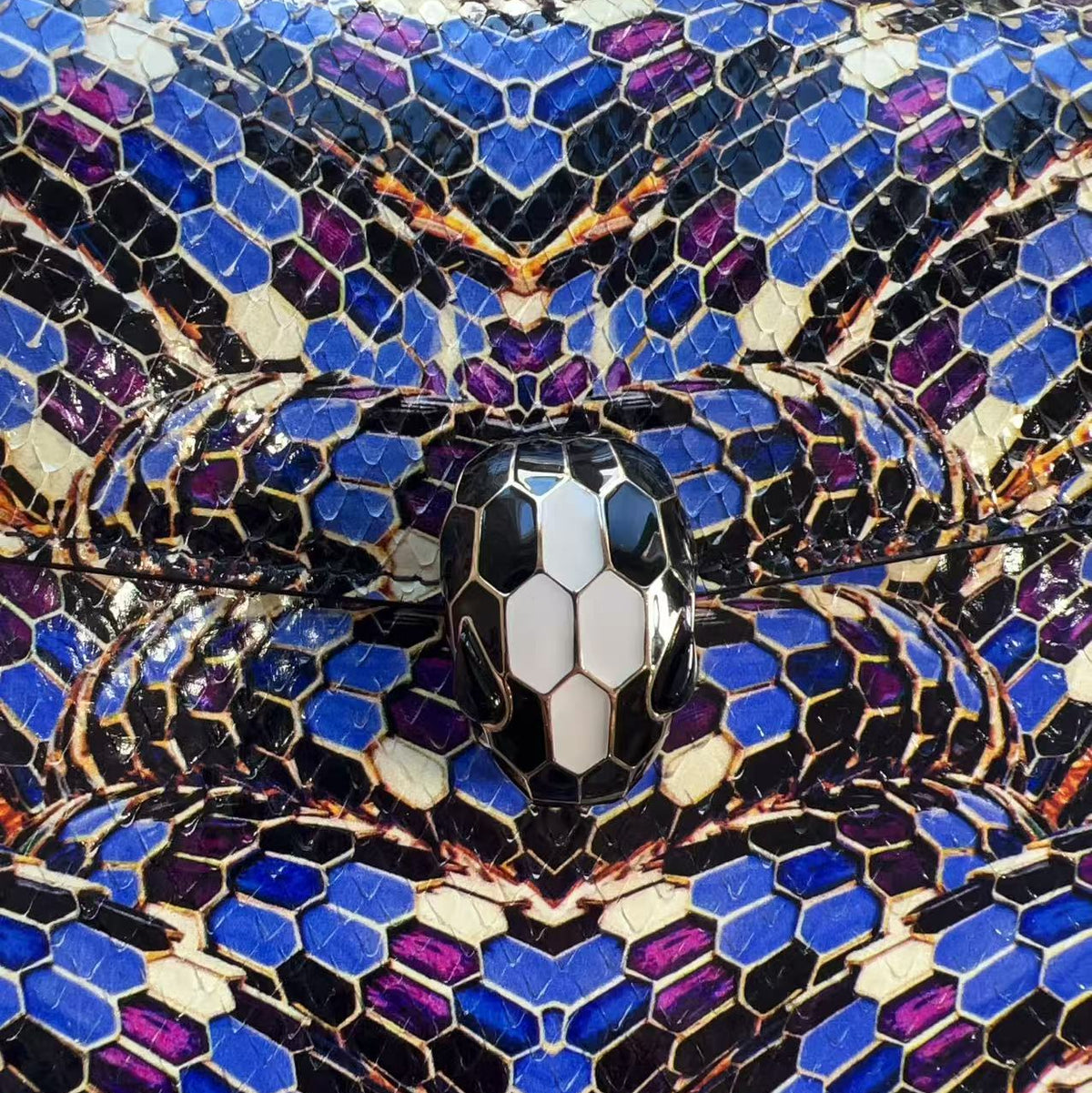 Close-up of a snakeskin-patterned handbag with blue, purple, black, and beige scales, featuring a central snake head clasp and a chain strap at the top.