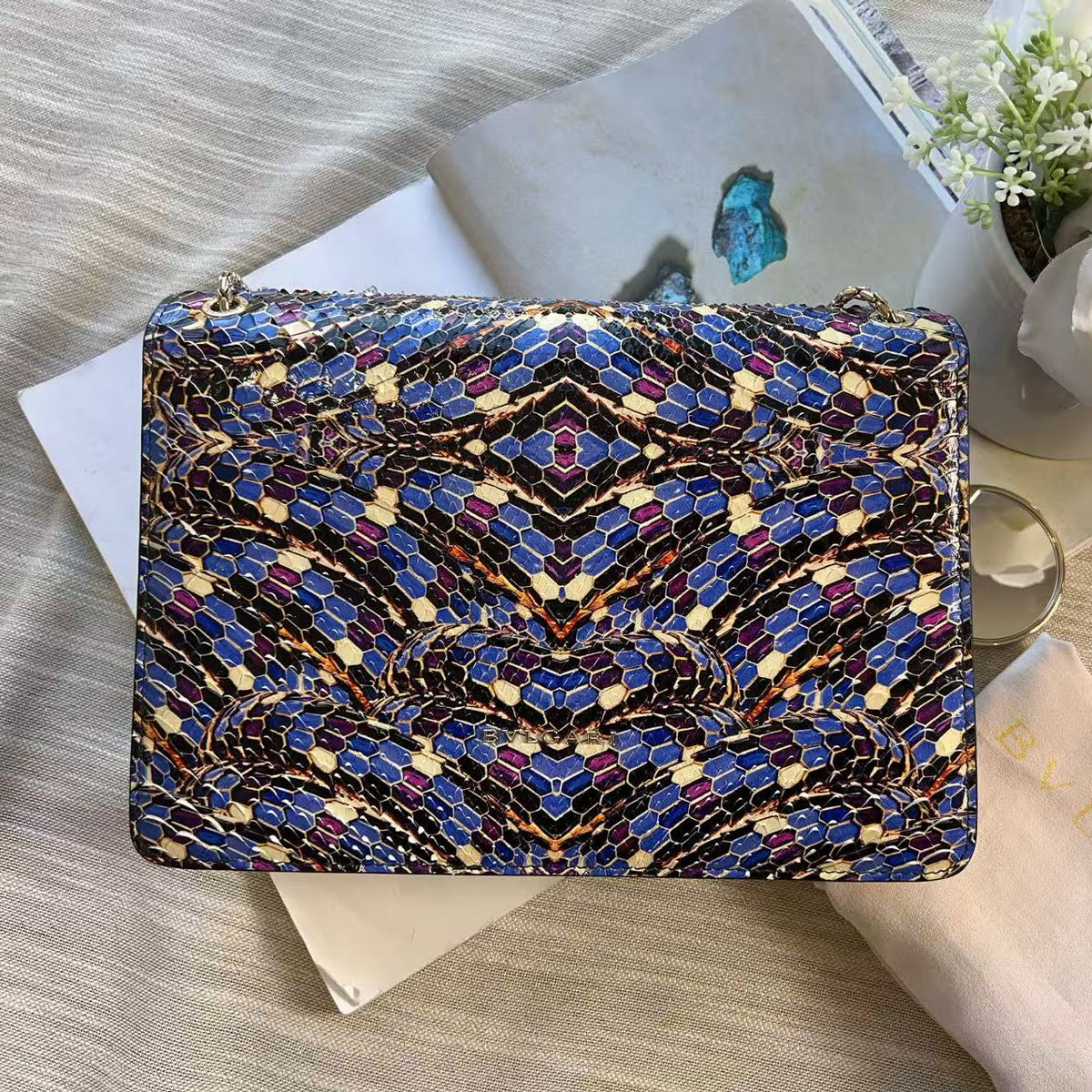 A rectangular zippered pouch or case with a blue, black, and gold snake-print pattern, styled on top of books and a beige satin bag with gold lettering.