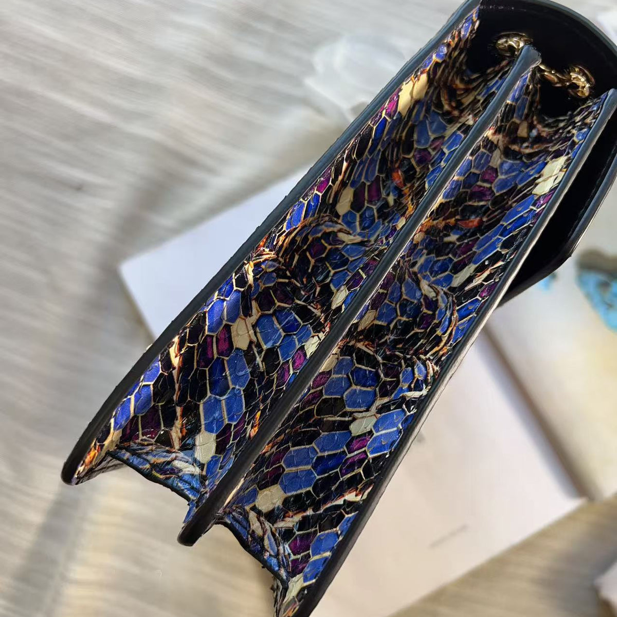 A close-up of a handbag's interior lining featuring a glossy, multicolored snakeskin pattern with blue, gold, black, and brown tones, bordered by black trim.