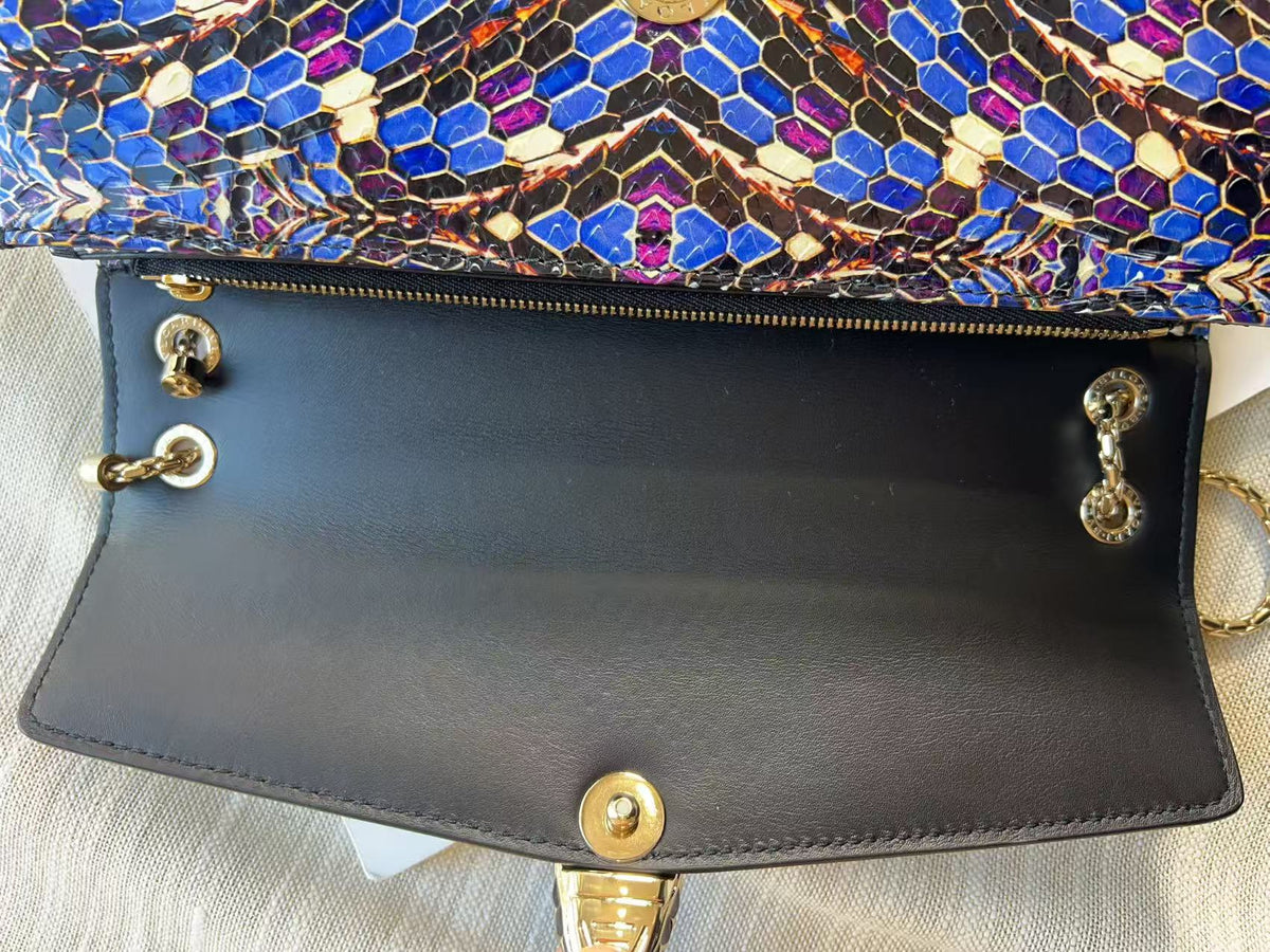 Close-up of a handbag showing the black leather interior flap with gold-tone hardware and a colorful snakeskin-patterned exterior in blue, purple, black, and gold.