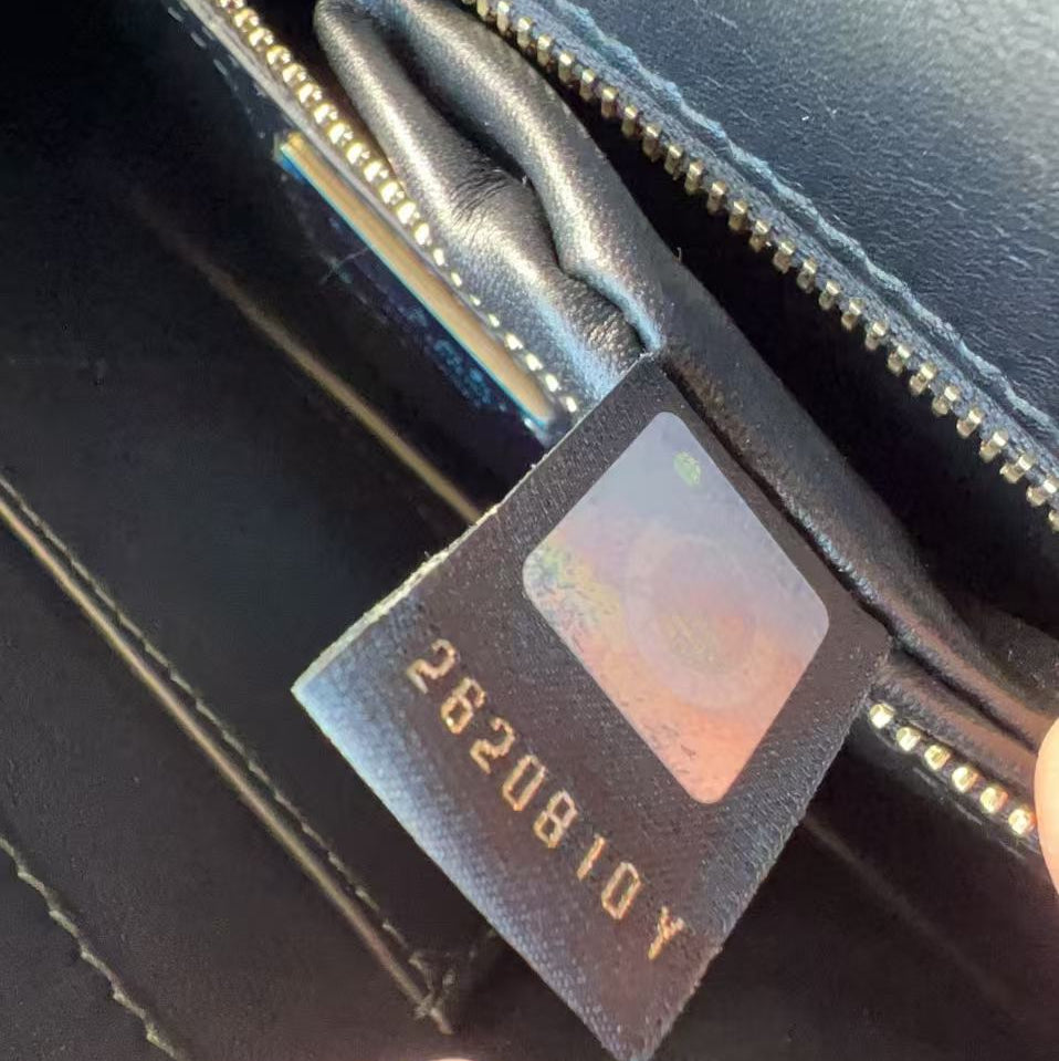 Close-up of a black leather zippered pouch or wallet interior, showing a sewn fabric tag with gold lettering and a holographic sticker, with fingers holding it open.