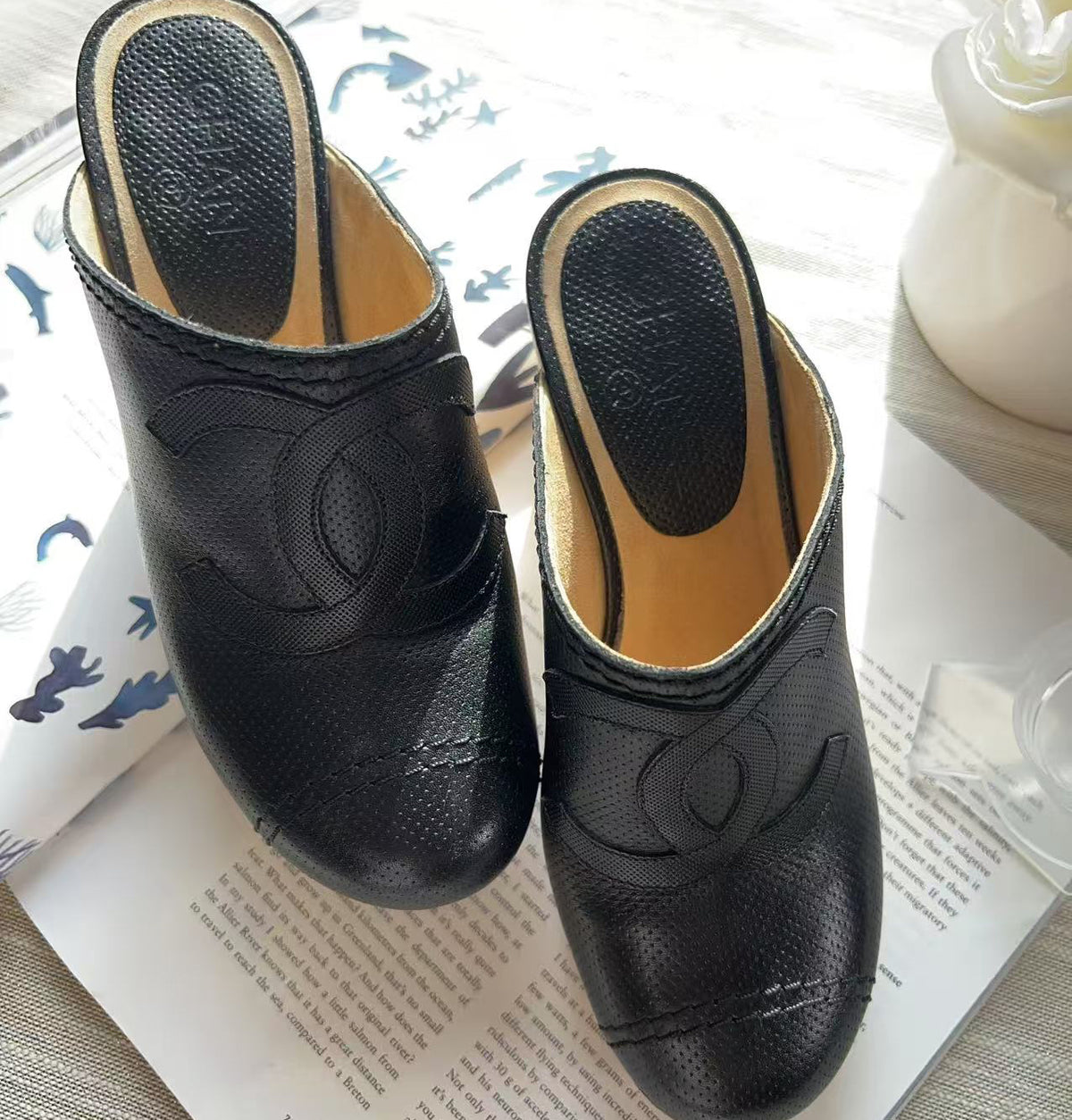 A pair of black leather mule slip-on shoes with a textured pebbled finish and a debossed interlocking CC logo on the side, resting on an open book.