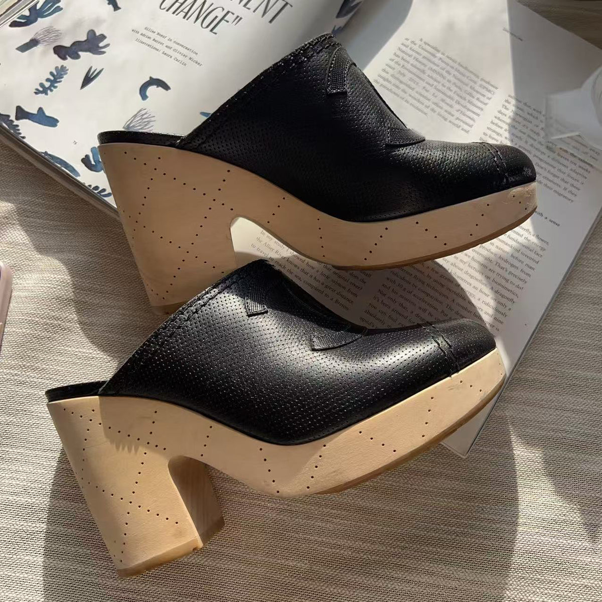 A pair of black pebbled leather mule clogs with chunky light beige cork-look platform heels, styled on top of open printed magazines on a light fabric surface.