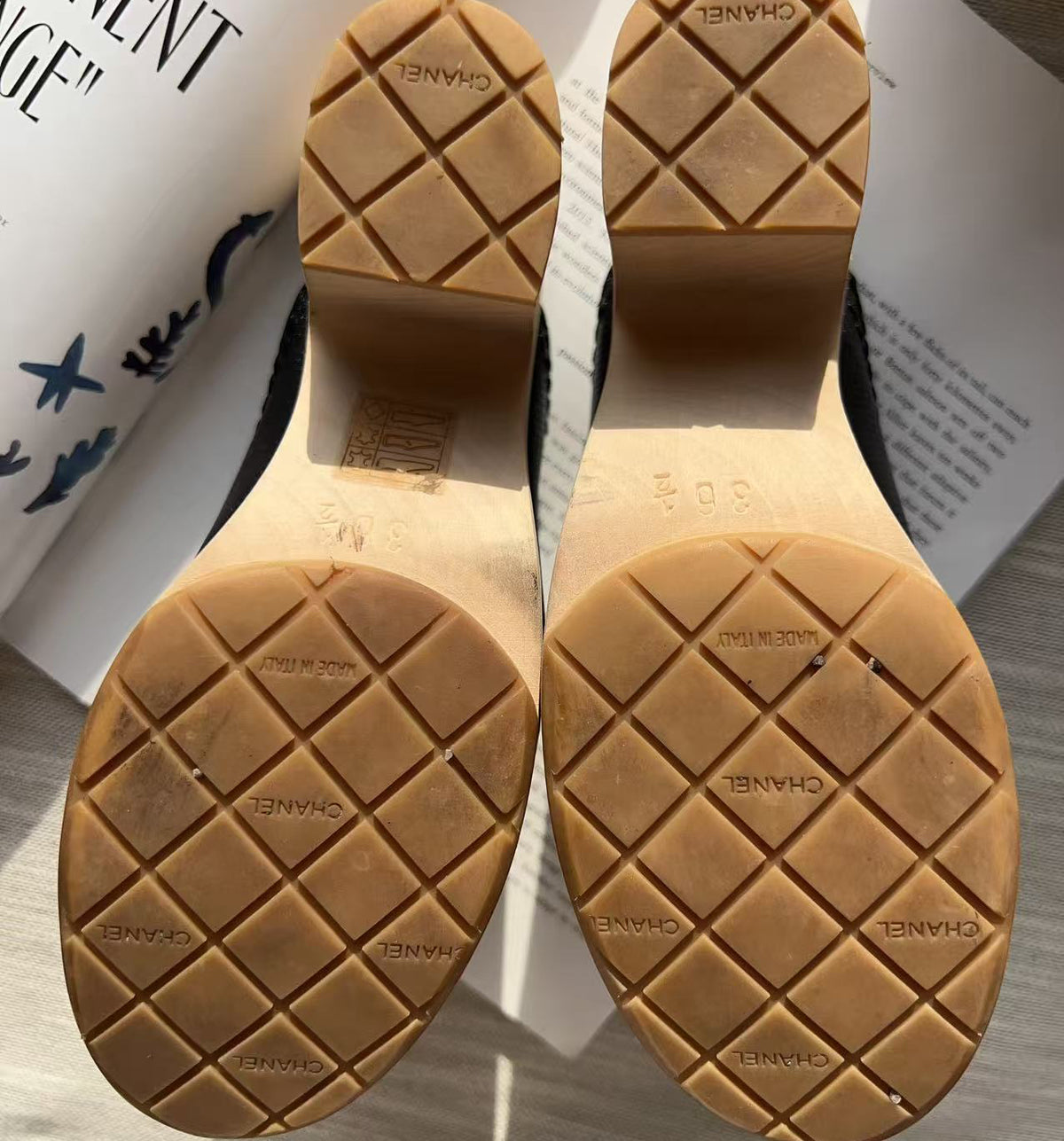 Close-up of the soles of a pair of Chanel high heels, showing the tan rubber outsoles with a diamond grid pattern and embossed CHANEL logos, photographed on a light wood surface with a magazine in the background.