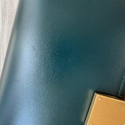 Close-up of a teal leather bag with a gold buckle on a textured beige surface.
