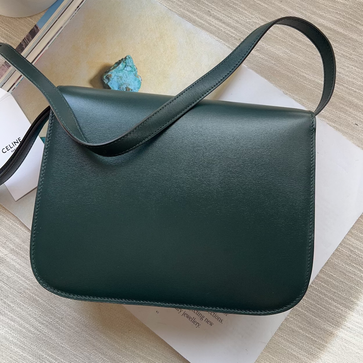 Green leather bag on a light wooden surface with a book and small plant.