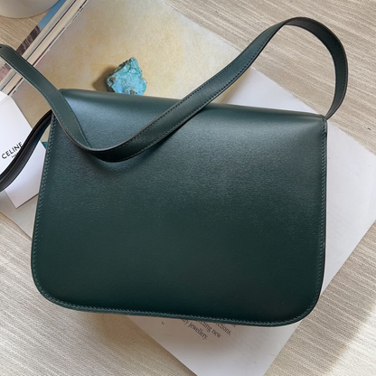 Green leather bag on a light wooden surface with a book and small plant.