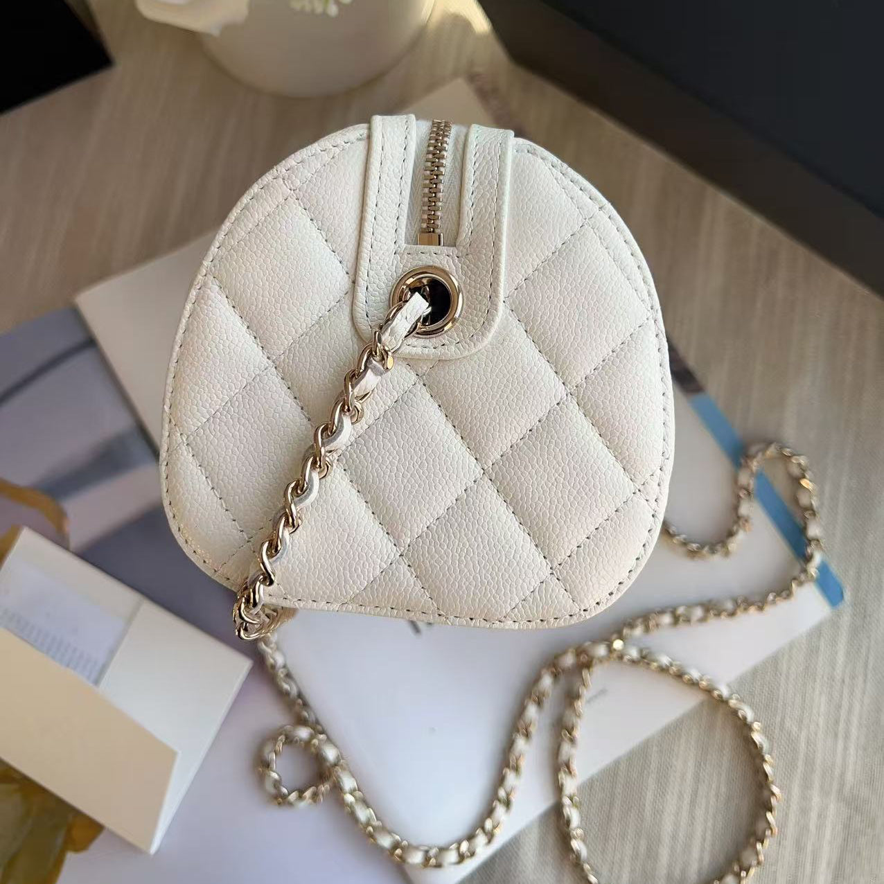 Quilted cream-colored handbag with gold chain on a light wooden surface with flowers and books.
