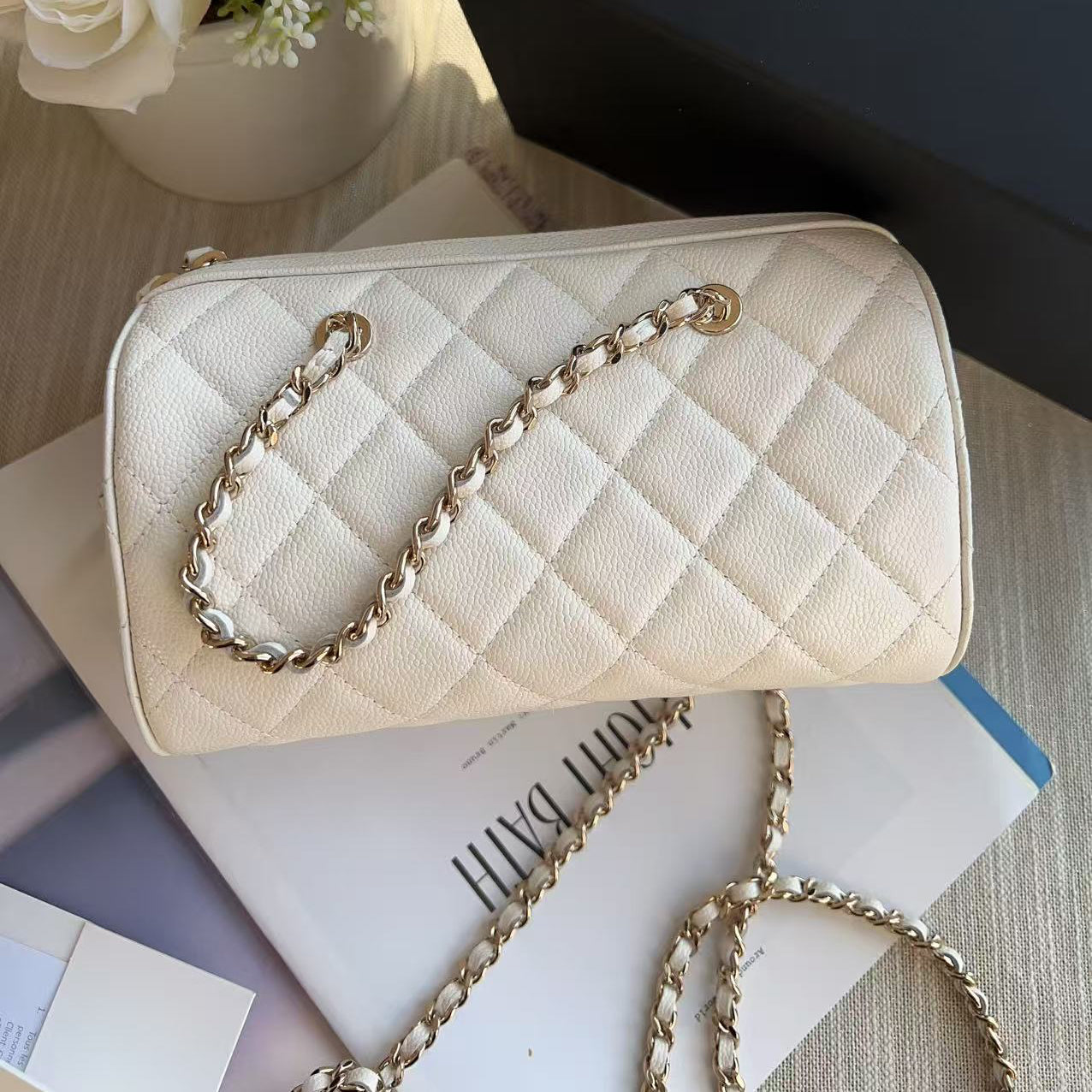 Beige quilted handbag with gold chain on a surface with flowers and books in the background