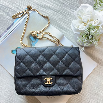 A black quilted leather handbag with a gold chain strap and a gold interlocking CC logo, styled on a light wood surface with a magazine and white flowers.