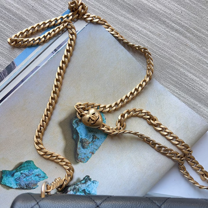 A gold-toned chunky chain necklace with a small sculpted pendant, displayed on top of a magazine page with turquoise stones, next to a black quilted leather bag.