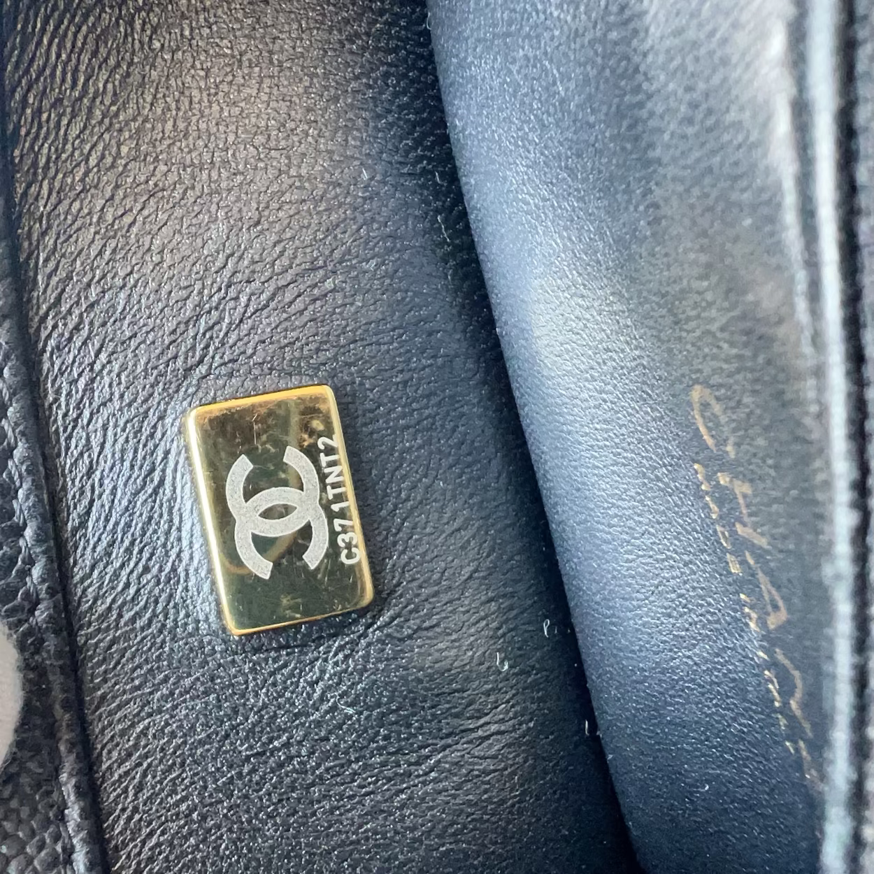 Close-up of a black leather Chanel bag showing a gold-tone metal plaque with the interlocking CC logo and 'CHANEL PARIS' text, with a white-gloved finger touching the leather.