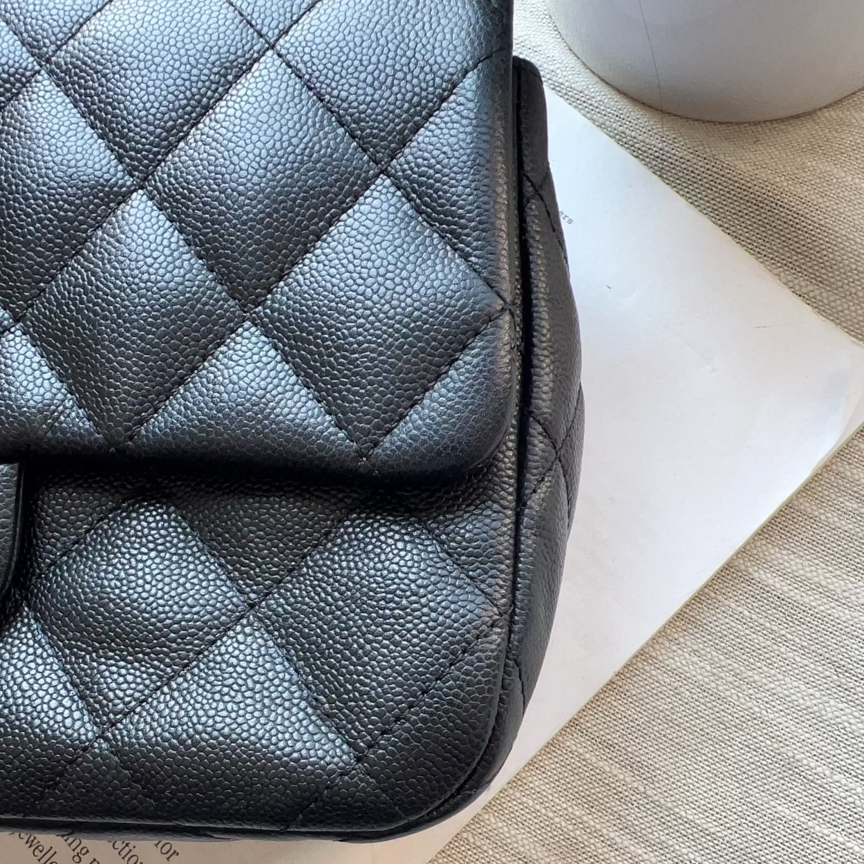 A close-up of a black quilted leather handbag with a pebbled texture, resting on a white sheet of paper over a beige woven surface.