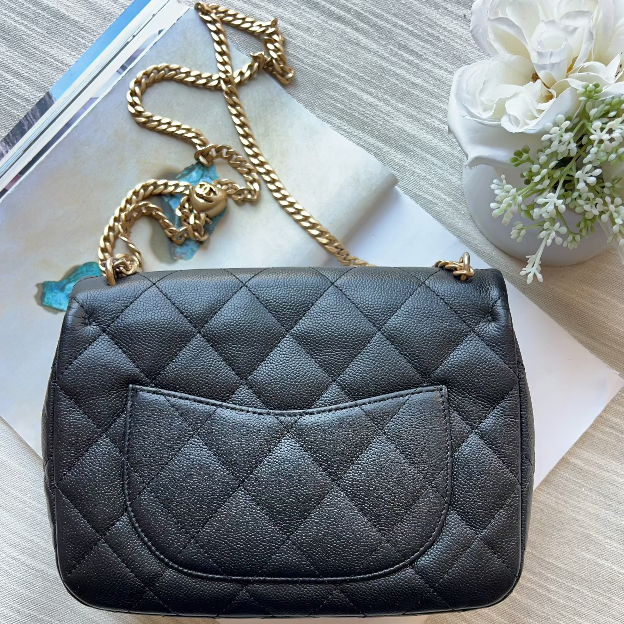 A black quilted leather handbag with a gold chain strap, styled on a light wood surface with a magazine, white paper, and a small white flower arrangement.