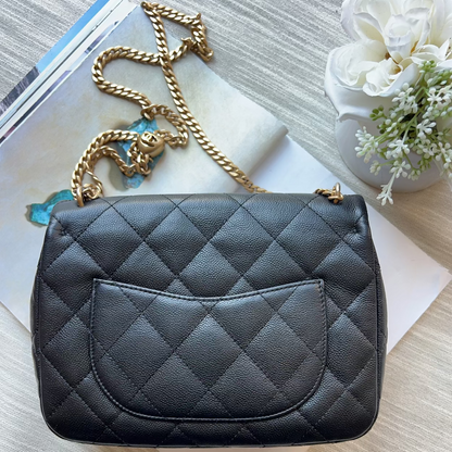 A black quilted leather handbag with a gold chain strap, styled on a light wood surface with a magazine, white paper, and a small white flower arrangement.