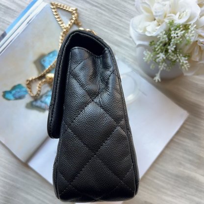 A black quilted leather handbag with a gold chain strap, shown from a top-down view resting on a white book over a light wood surface with decorative flowers in the background.