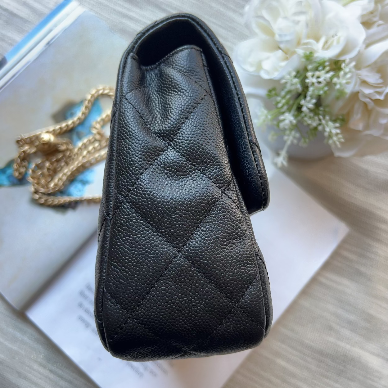 A black quilted leather handbag with a pebbled texture, shown from an overhead angle resting on a light wood surface with a gold chain, an open book, and white flowers in the background.