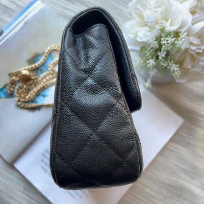 A black quilted leather handbag with a pebbled texture, shown from an overhead angle resting on a light wood surface with a gold chain, an open book, and white flowers in the background.