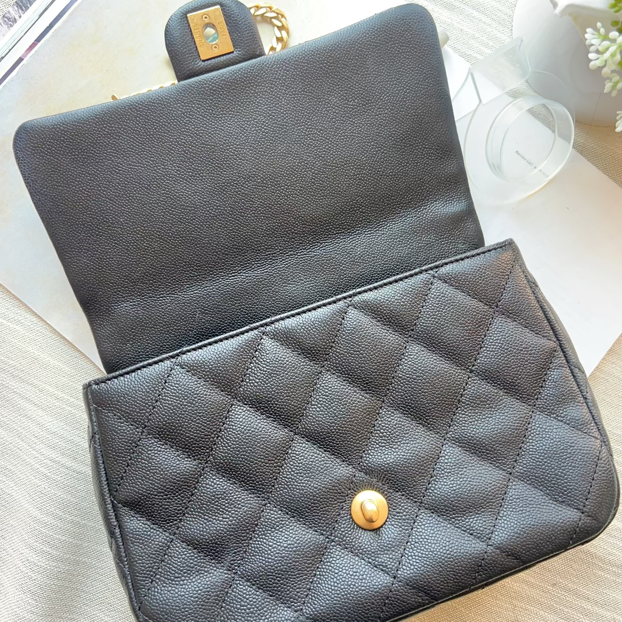 A black quilted leather handbag with a gold snap button and a gold-tone clasp, shown open on a light wood surface with a magazine and white vase in the background.