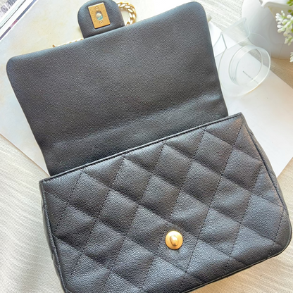 A black quilted leather handbag with a gold snap button and a gold-tone clasp, shown open on a light wood surface with a magazine and white vase in the background.