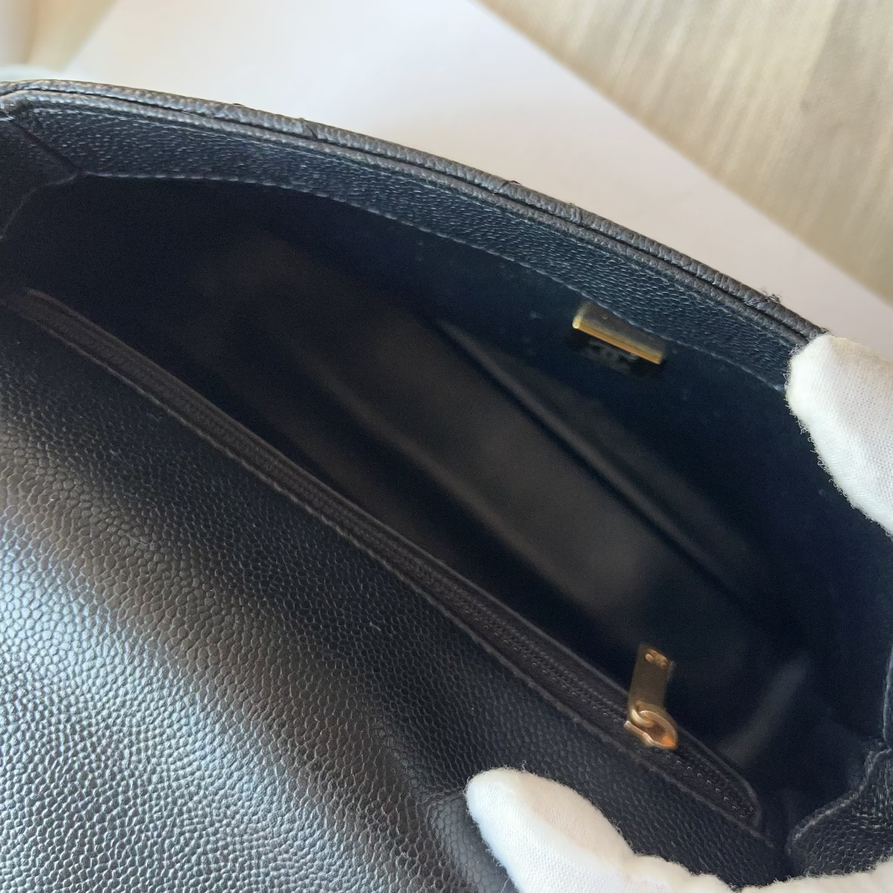 Close-up of a black pebbled leather clutch or handbag interior with a white-gloved hand holding it open, showing a black lining and small gold-tone hardware.