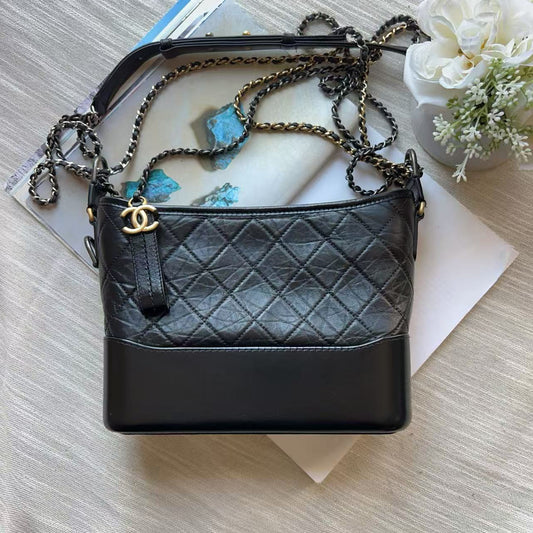 A black quilted leather shoulder bag with a gold-tone interlocking CC logo and a chain strap, styled on top of books and a small white flower arrangement.