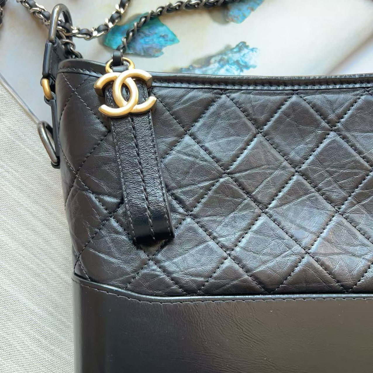 Close-up of a black quilted leather handbag with a gold-tone interlocking CC logo charm and chain strap, resting on a light textured surface.