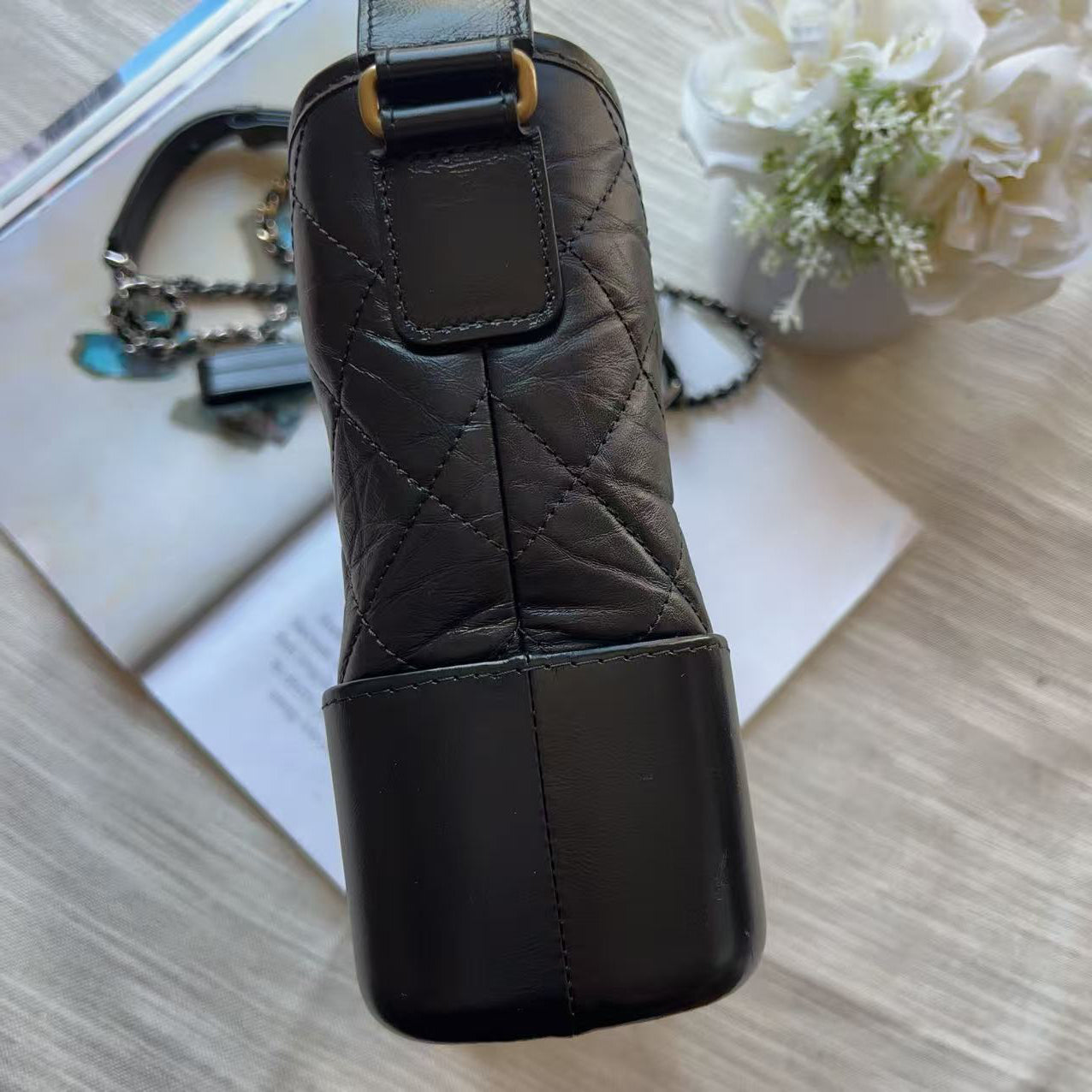 A close-up of a black quilted leather handbag or clutch with a flap closure and gold-tone hardware, photographed from above on a light wood surface with a book and decorative flowers in the background.