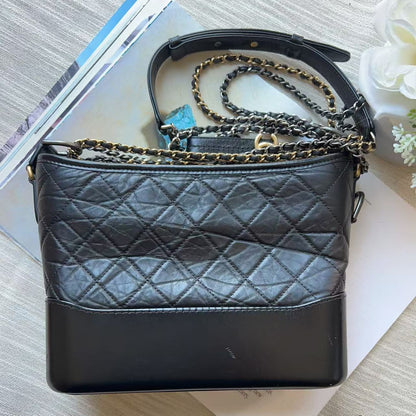 A black quilted leather shoulder bag with a smooth black leather side panel and a gold-tone chain strap, styled on top of books on a light wood surface.