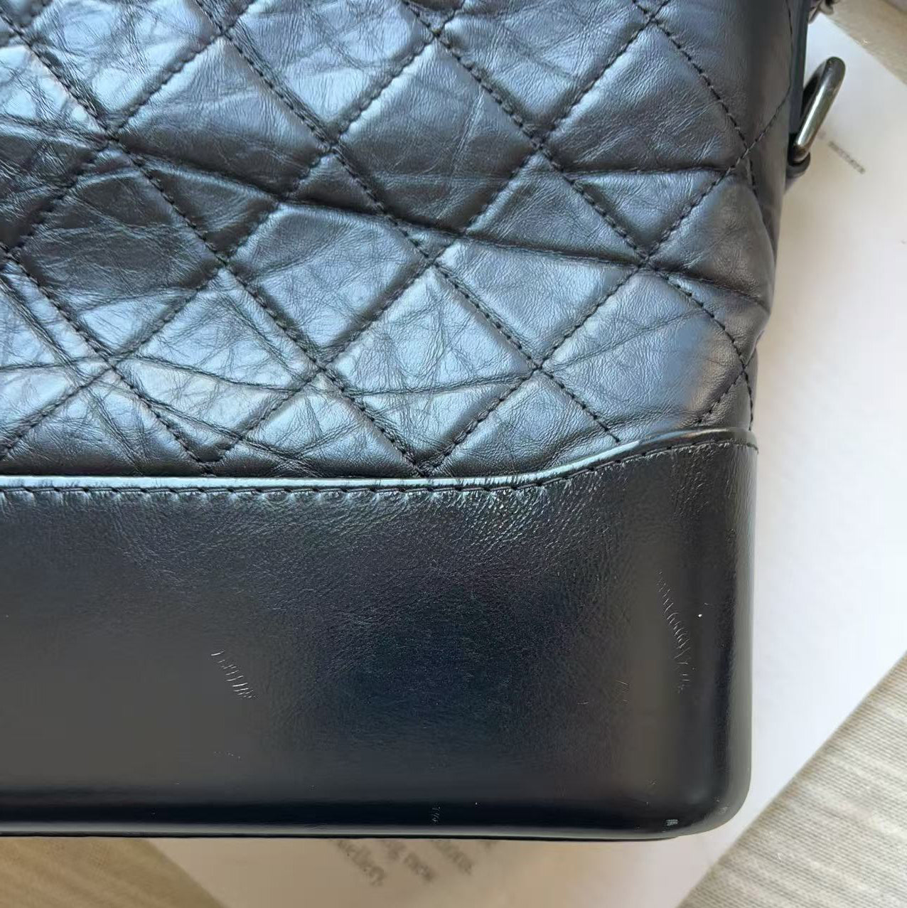 Close-up of a black quilted leather handbag corner with diamond stitching, placed on a white surface over a light wood floor.