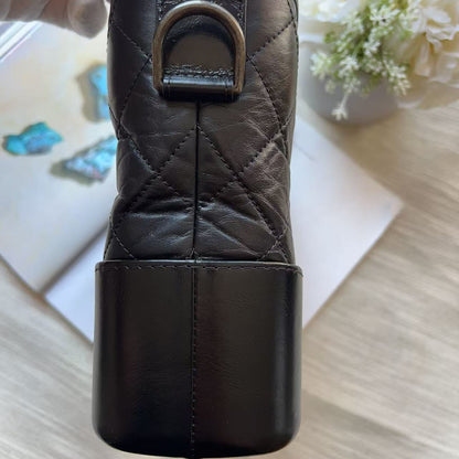 A close-up of a dark brown quilted leather accessory, likely a pouch or case, held by a hand over a light wood surface with a white flower and book in the background.