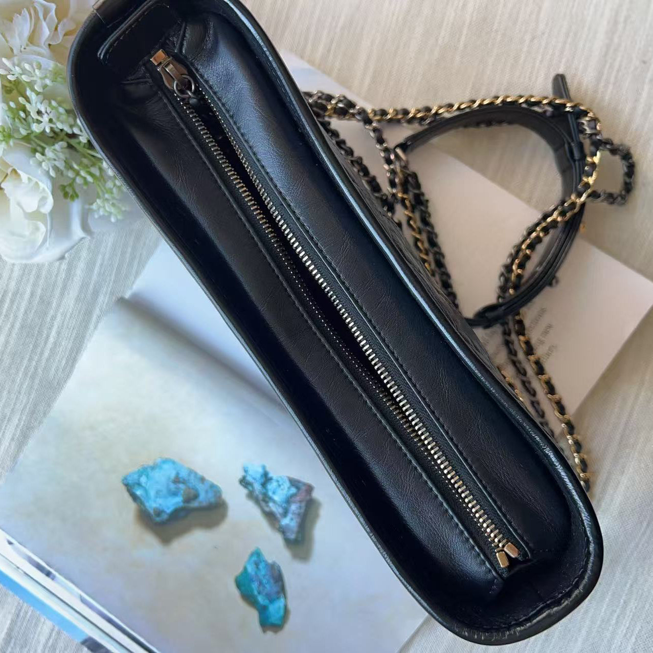 Black leather zippered clutch or pencil case with a gold-tone zipper and chain strap, styled on top of an open book with white flowers and turquoise stones nearby.