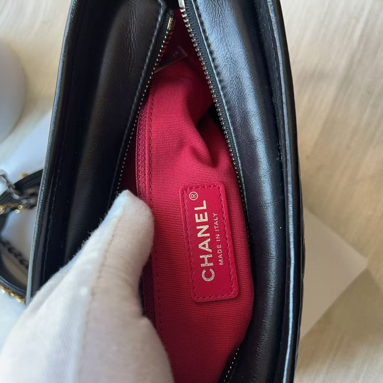 Close-up of a black leather Chanel handbag interior with a bright pink-red lining and a visible Chanel Made in Italy label, shown with a white gloved hand inside.