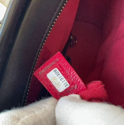 Close-up of a black leather handbag interior with a bright red lining and a red fabric tag being held by a white-gloved hand. A portion of a black-and-gold chain strap is visible on the left.