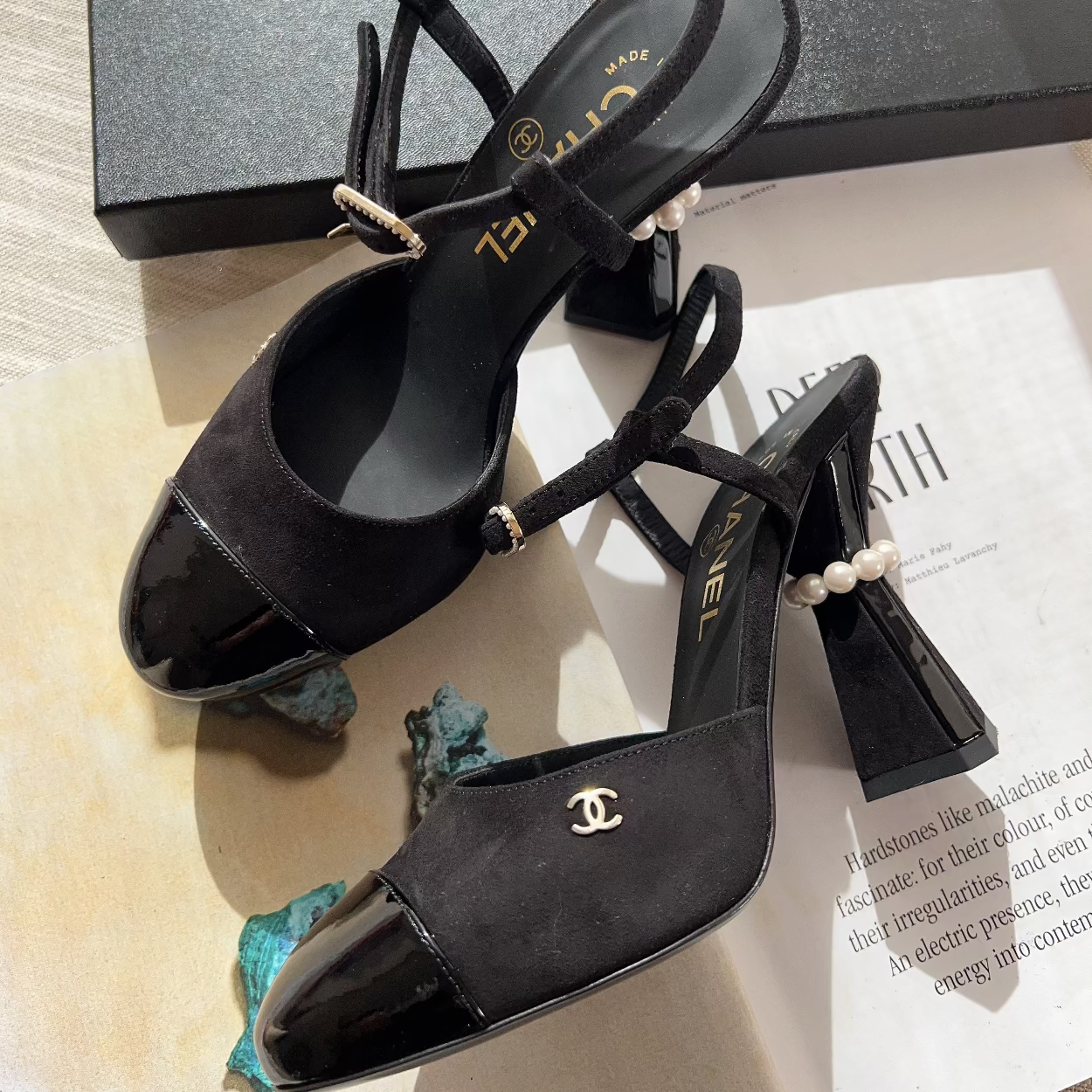 Black high-heeled sandals with pearl details on a surface with a Chanel box in the background.