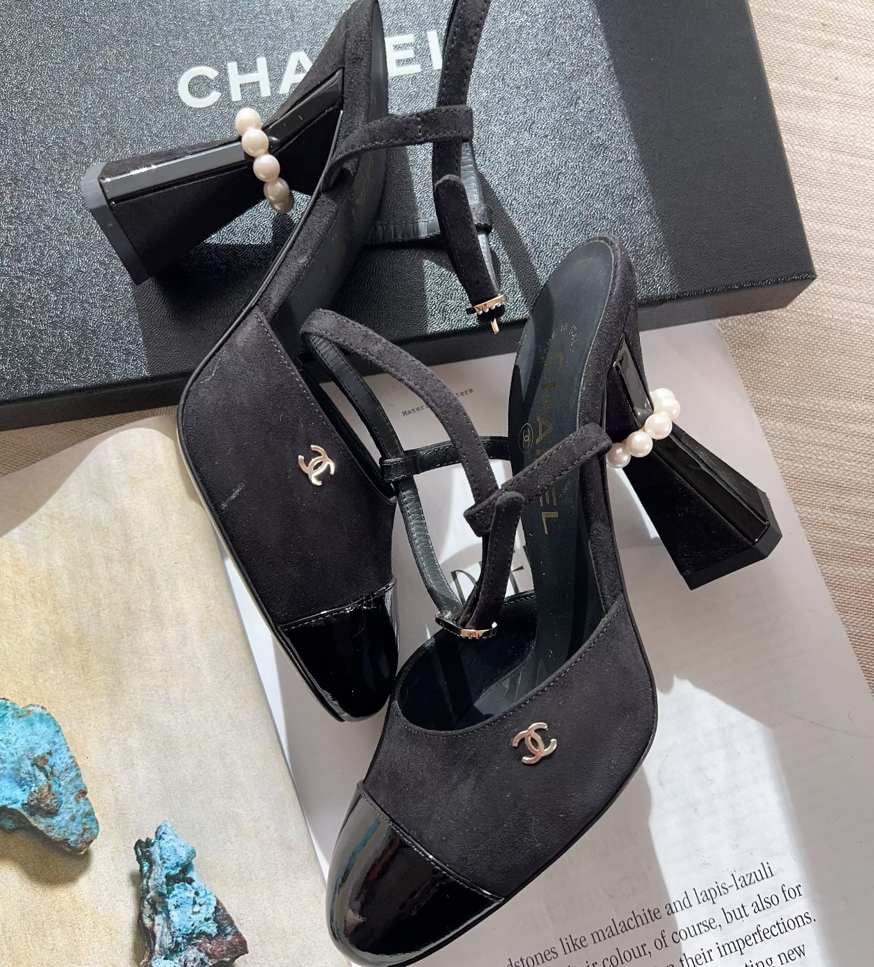 Black high-heeled shoes with pearl details on a surface with a Chanel box in the background.