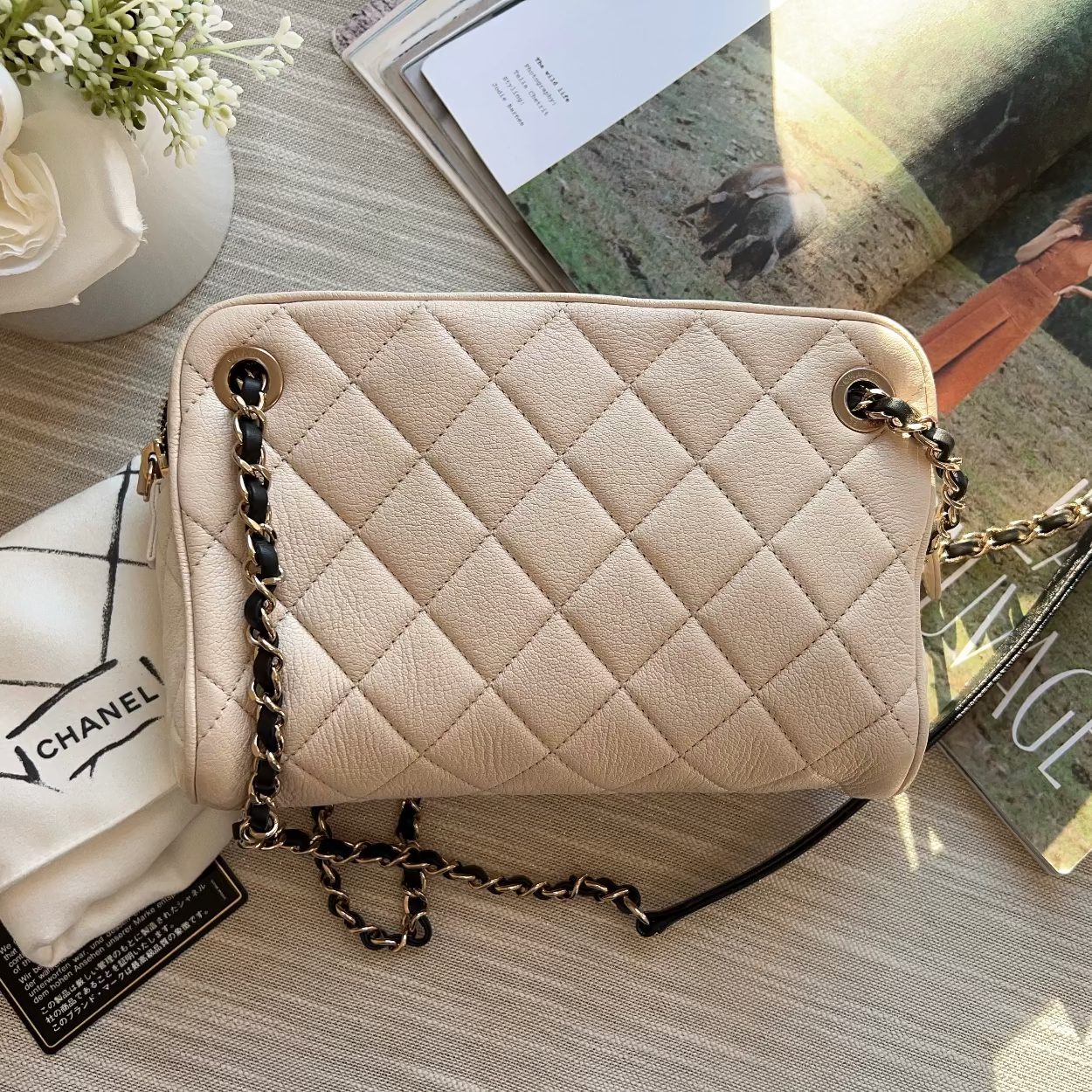 Beige quilted handbag with gold chain on a wooden surface with a Chanel sweatshirt and magazine.