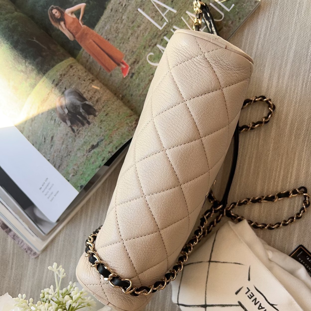 Beige quilted handbag with gold chain on a surface with a magazine and flowers.