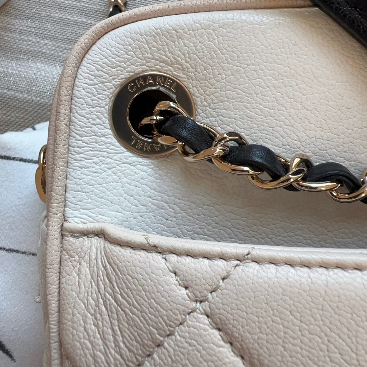 Close-up of a beige Chanel handbag with a gold chain strap on a textured surface.