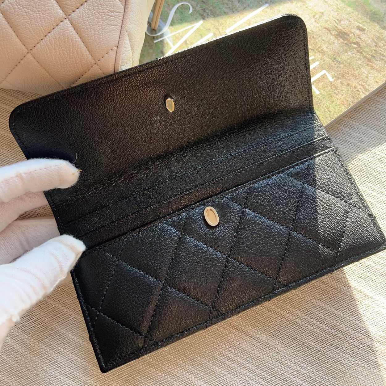 Black quilted wallet held by a gloved hand with a beige quilted handbag in the background.