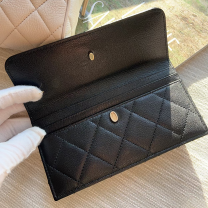 Black quilted wallet held by a gloved hand with a beige quilted handbag in the background.