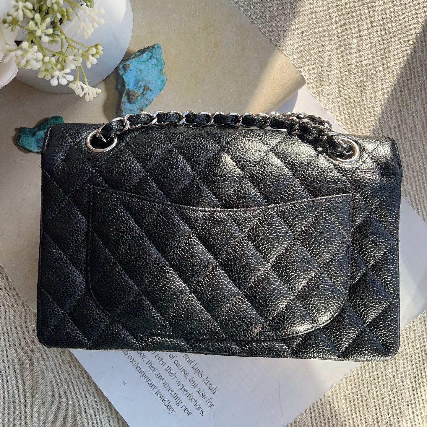 Black quilted handbag with chain detail on a textured surface with flowers and a book.