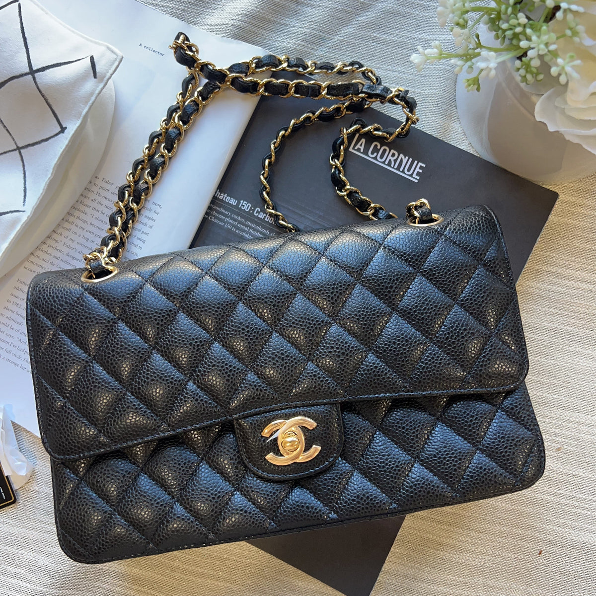 A black quilted leather shoulder bag with gold-tone chain straps and a gold interlocking CC logo, styled on top of a black book over a light beige textured surface.