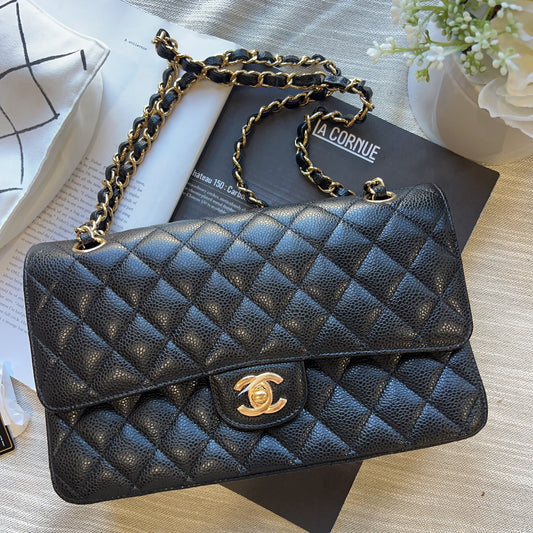 A black quilted leather shoulder bag with gold-tone chain straps and a gold interlocking CC logo, styled on top of a black book over a light beige textured surface.