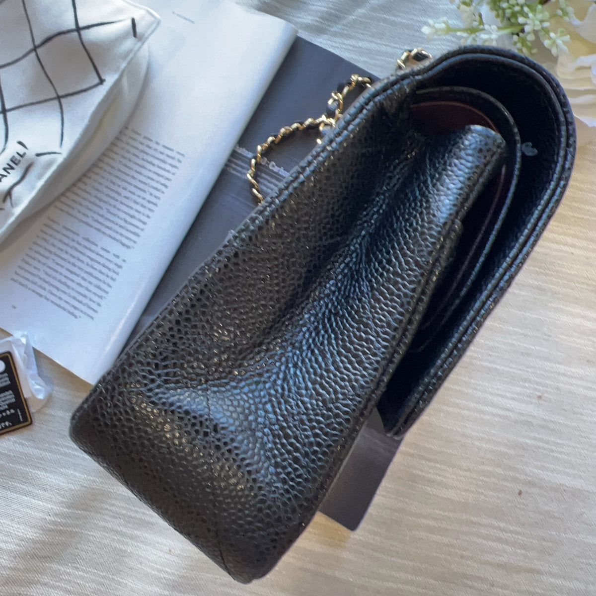 A black pebbled leather handbag with a gold chain strap, photographed on a light wood surface next to an open book and a white bag.