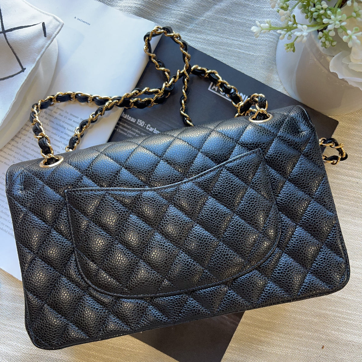 A black quilted caviar leather handbag with gold-tone chain strap details, styled on top of a black book over a light wood surface.