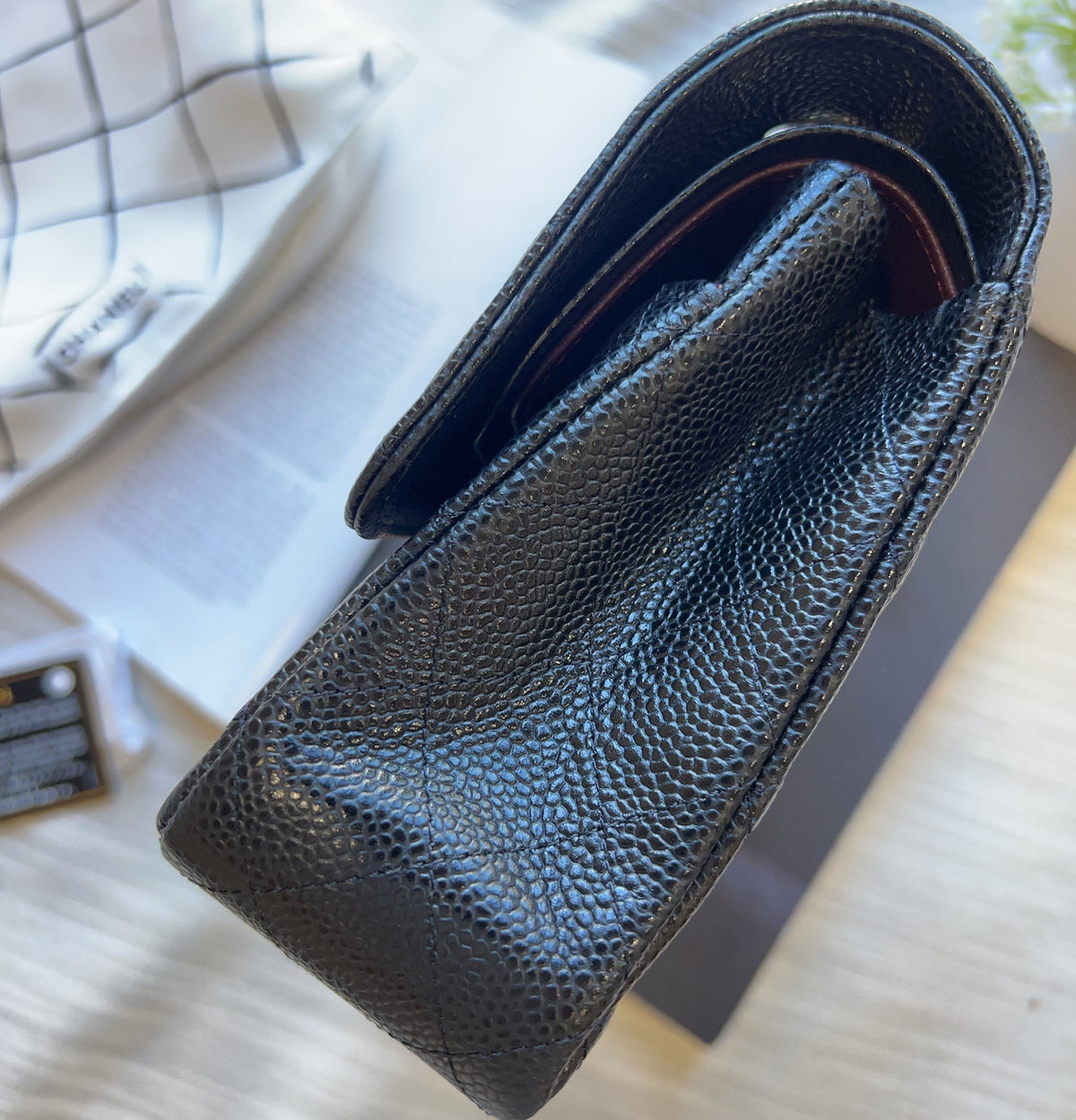 A close-up of a black pebbled leather pouch or case, showing the textured grain and curved shape, resting on a light wood surface next to a white checkered cloth.