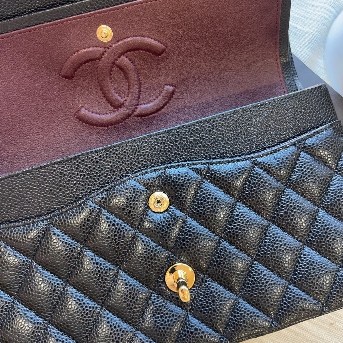 Close-up of a luxury handbag interior showing a black pebbled leather quilted flap with gold-tone hardware and a burgundy leather interior with an embossed interlocking CC logo, indicating Chanel.